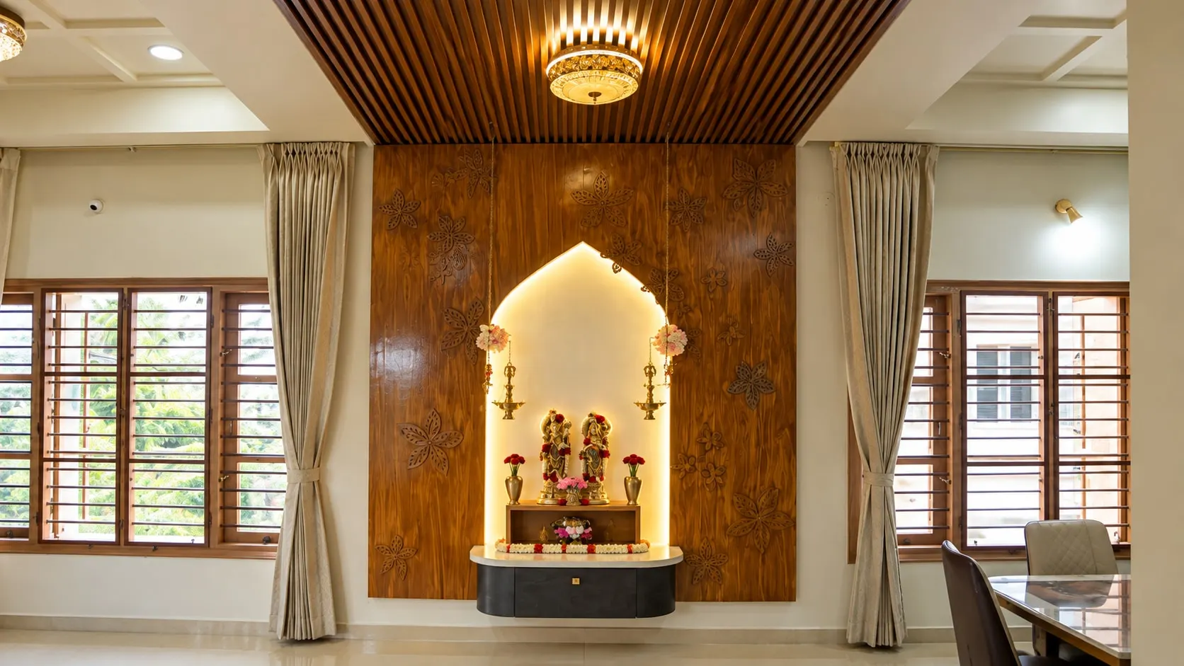 Pooja wall of carved timber panels with a lit arched niche, under a timber-slat ceiling, between two windows