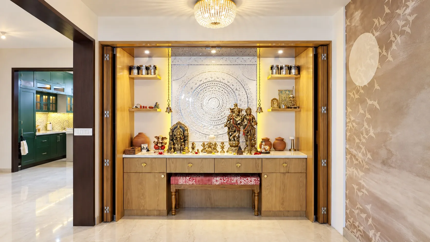 Pooja room with a carved white panel, brass idols, lit timber shelves and folding timber doors, with the green kitchen beyond