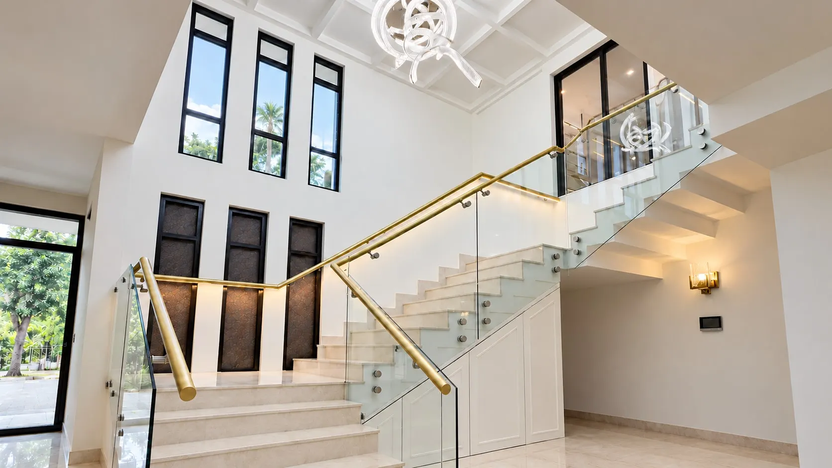Double-height staircase with a brass handrail and glass balustrade, white marble steps and tall black-framed windows