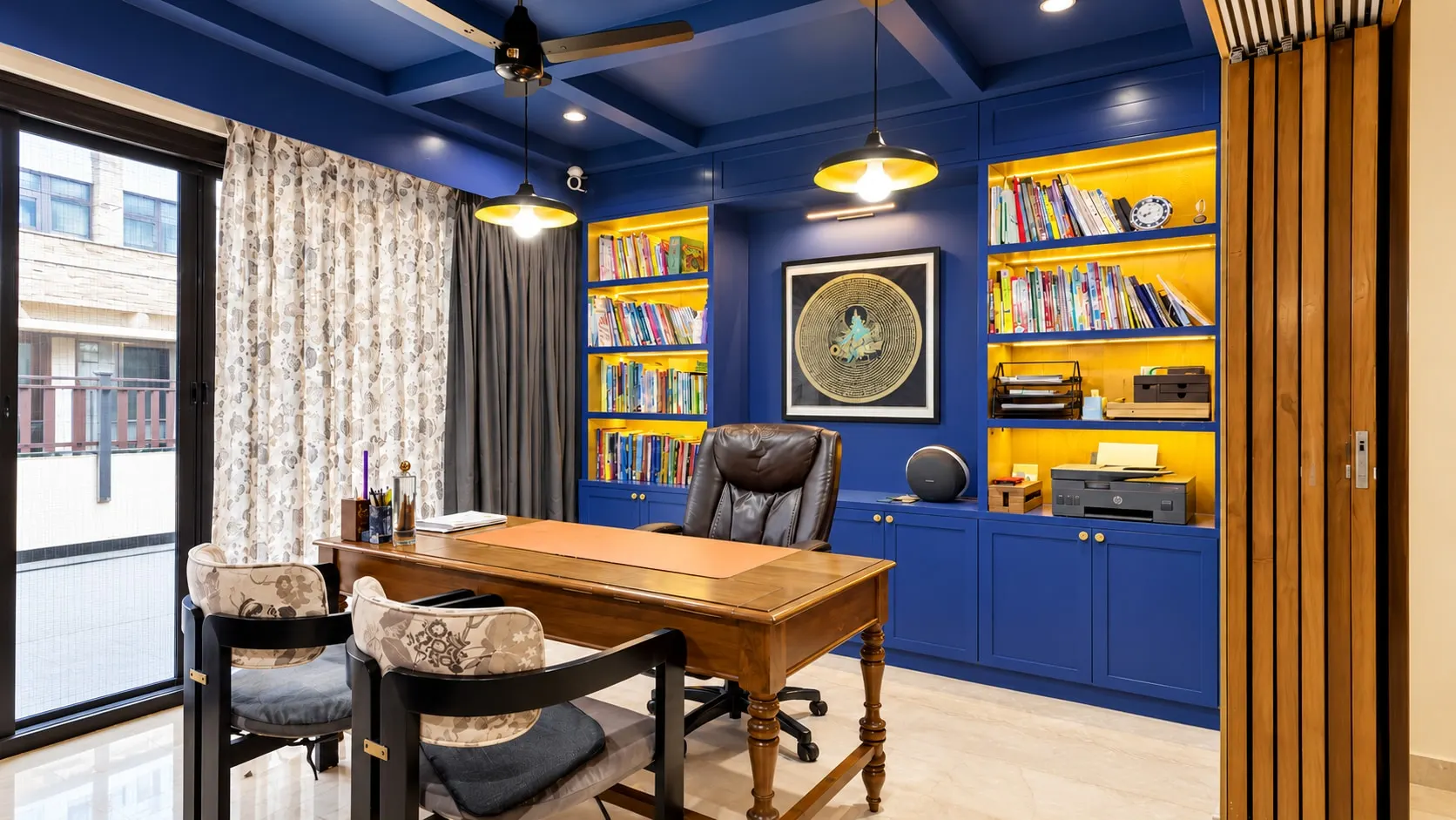 Study with cobalt-blue bookshelves lit in warm yellow, a coffered blue ceiling, a timber desk and two pendant lights