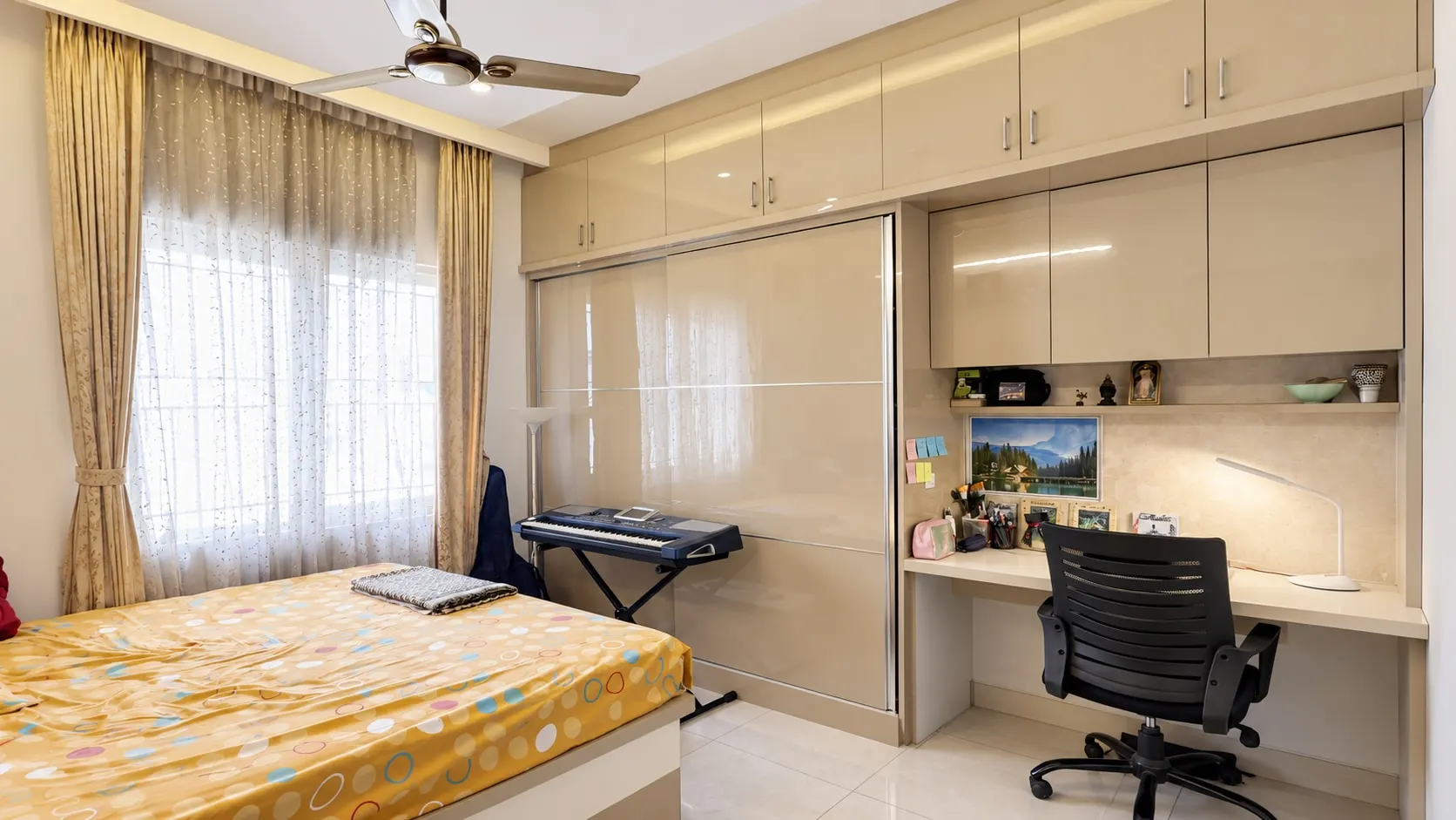 Bedroom with a cream gloss wardrobe that runs into a study desk with open shelves, a keyboard on a stand and curtained windows