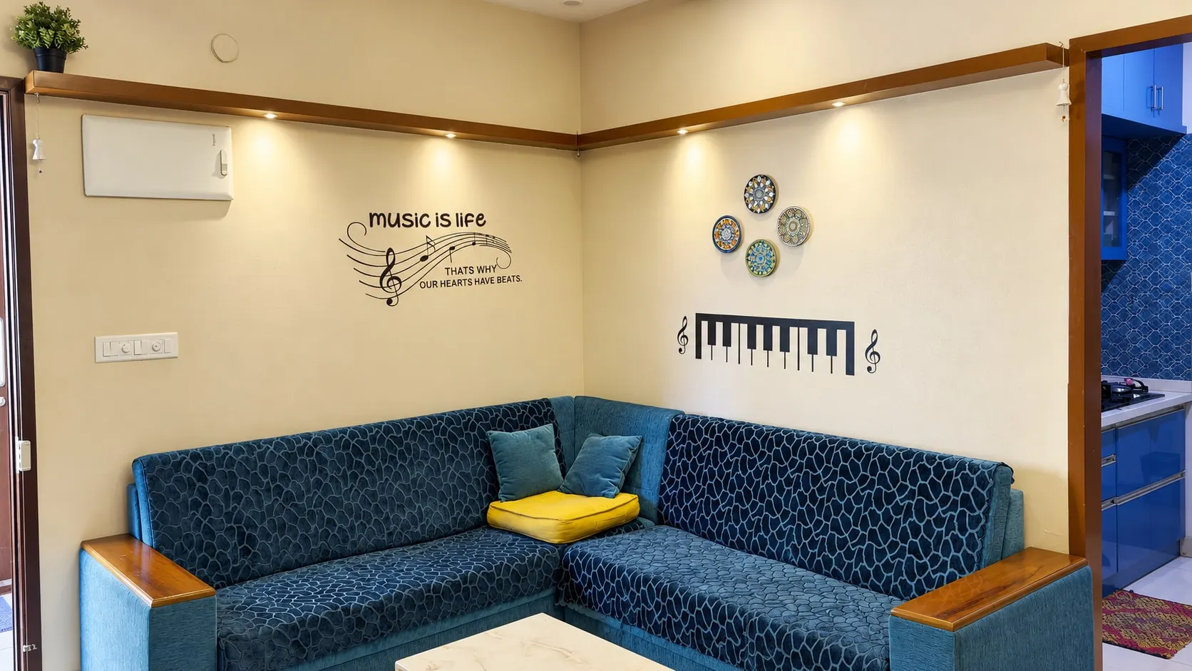 Blue corner sofa under a wall that reads “music is life” beside a row of piano keys, with the blue kitchen through the doorway