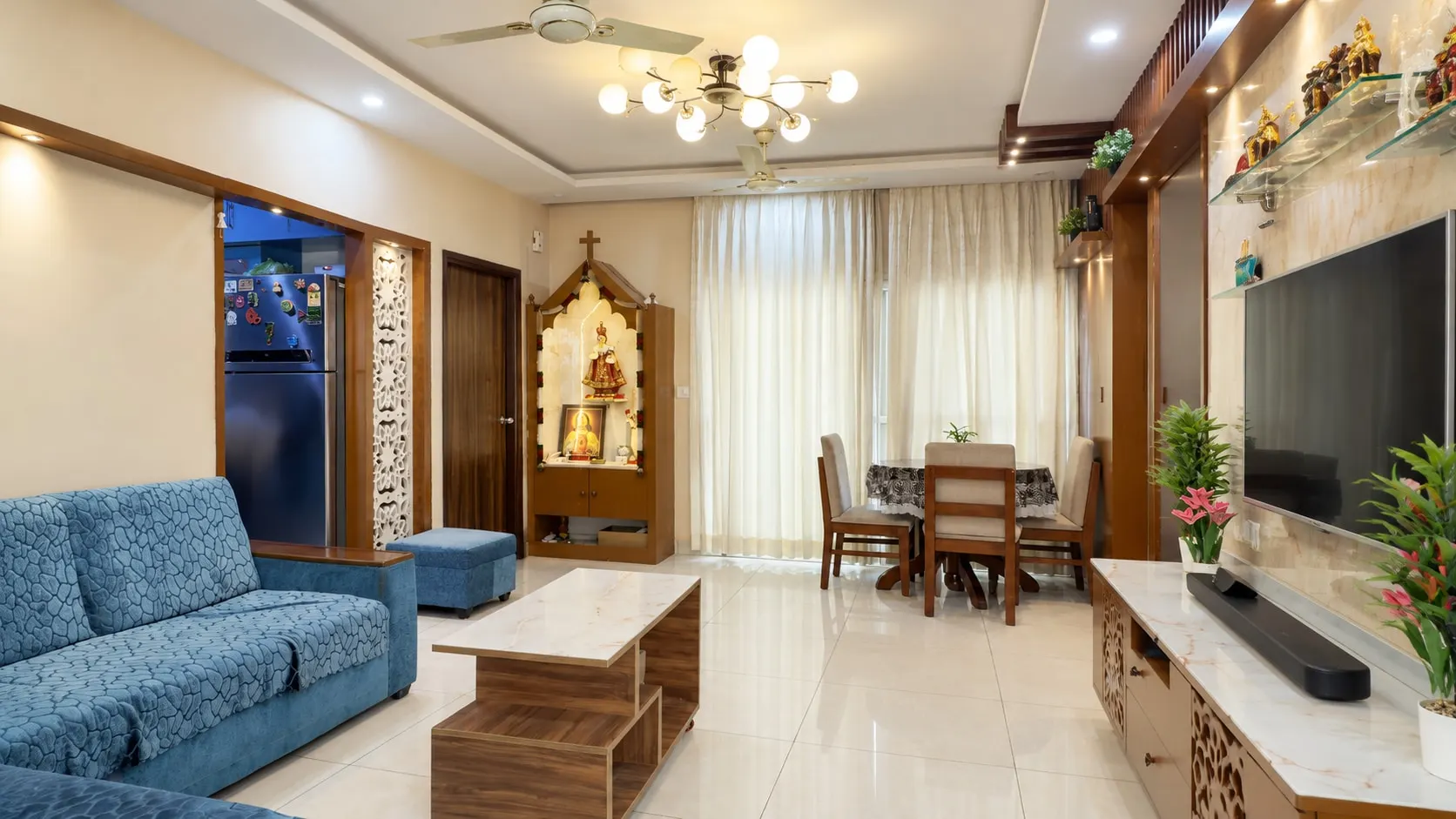 Living and dining room with a blue sofa, a marble-top coffee table, a timber prayer unit, a marble-finish TV wall with glass shelves and a round dining table by the window