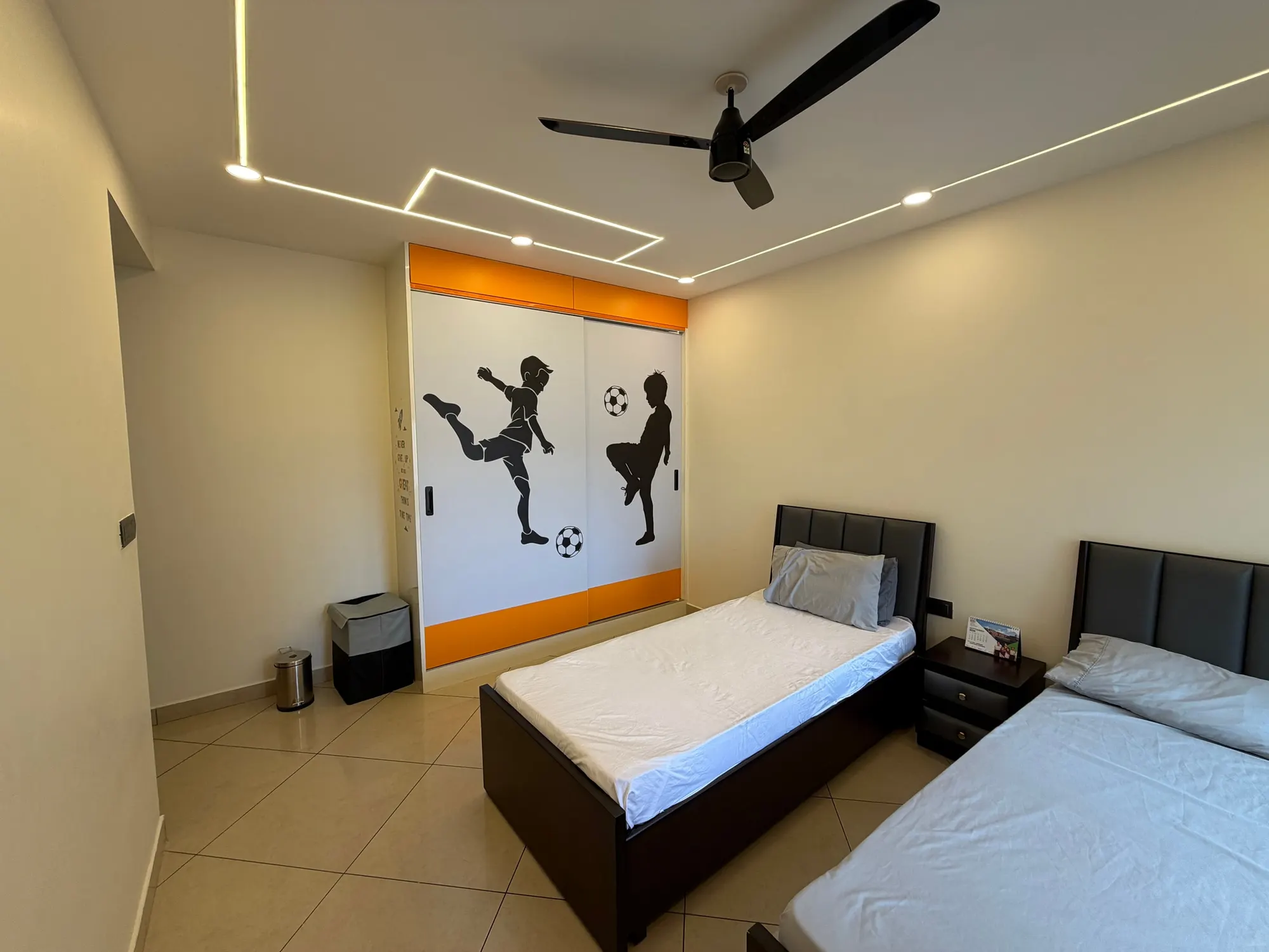 Bedroom with twin beds, a sliding wardrobe printed with footballers in orange and white, and lines of light set into the ceiling
