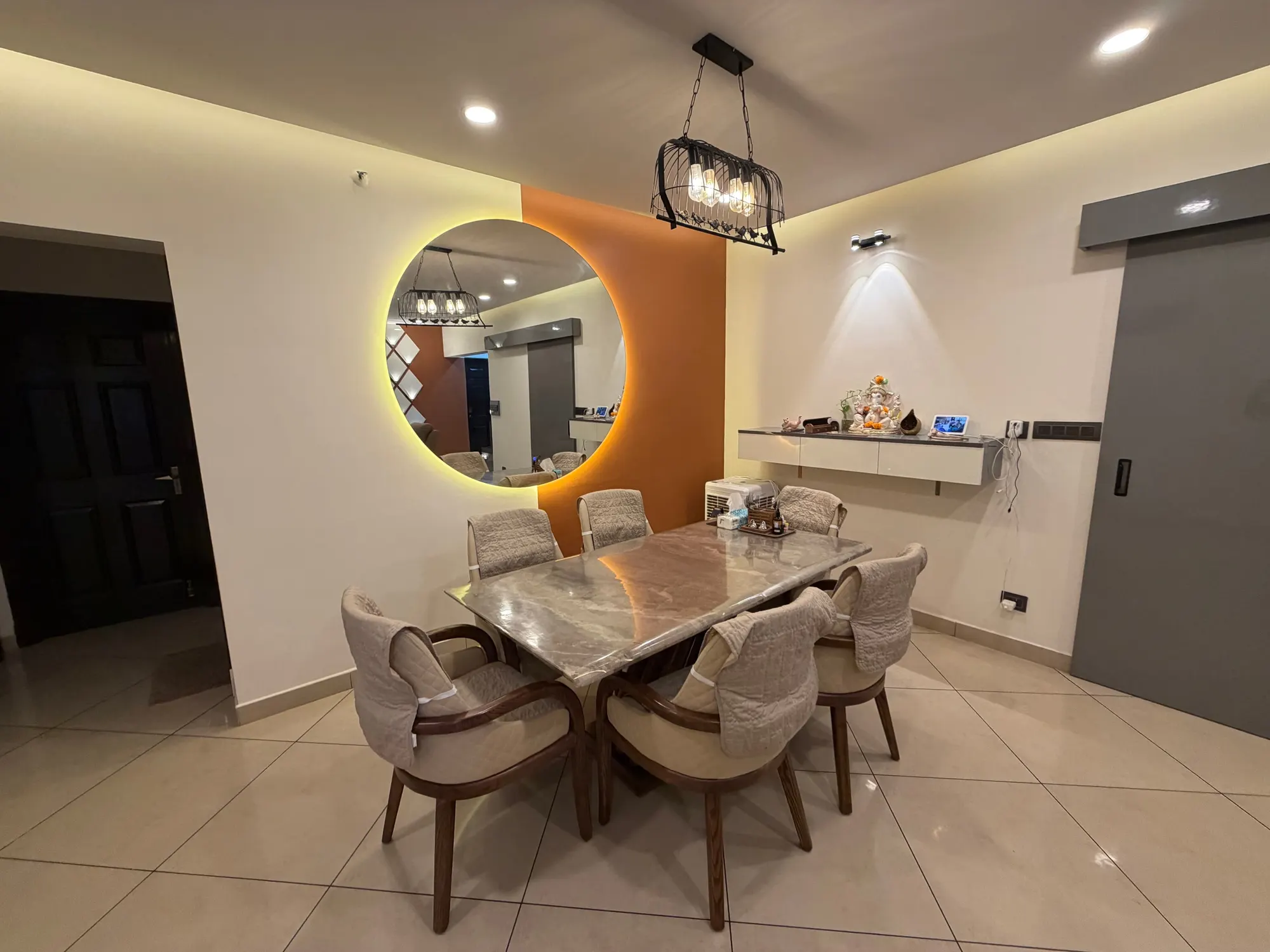 Dining table with a marble top and curved timber chairs under a cage pendant, beside a backlit round mirror on a burnt-orange wall
