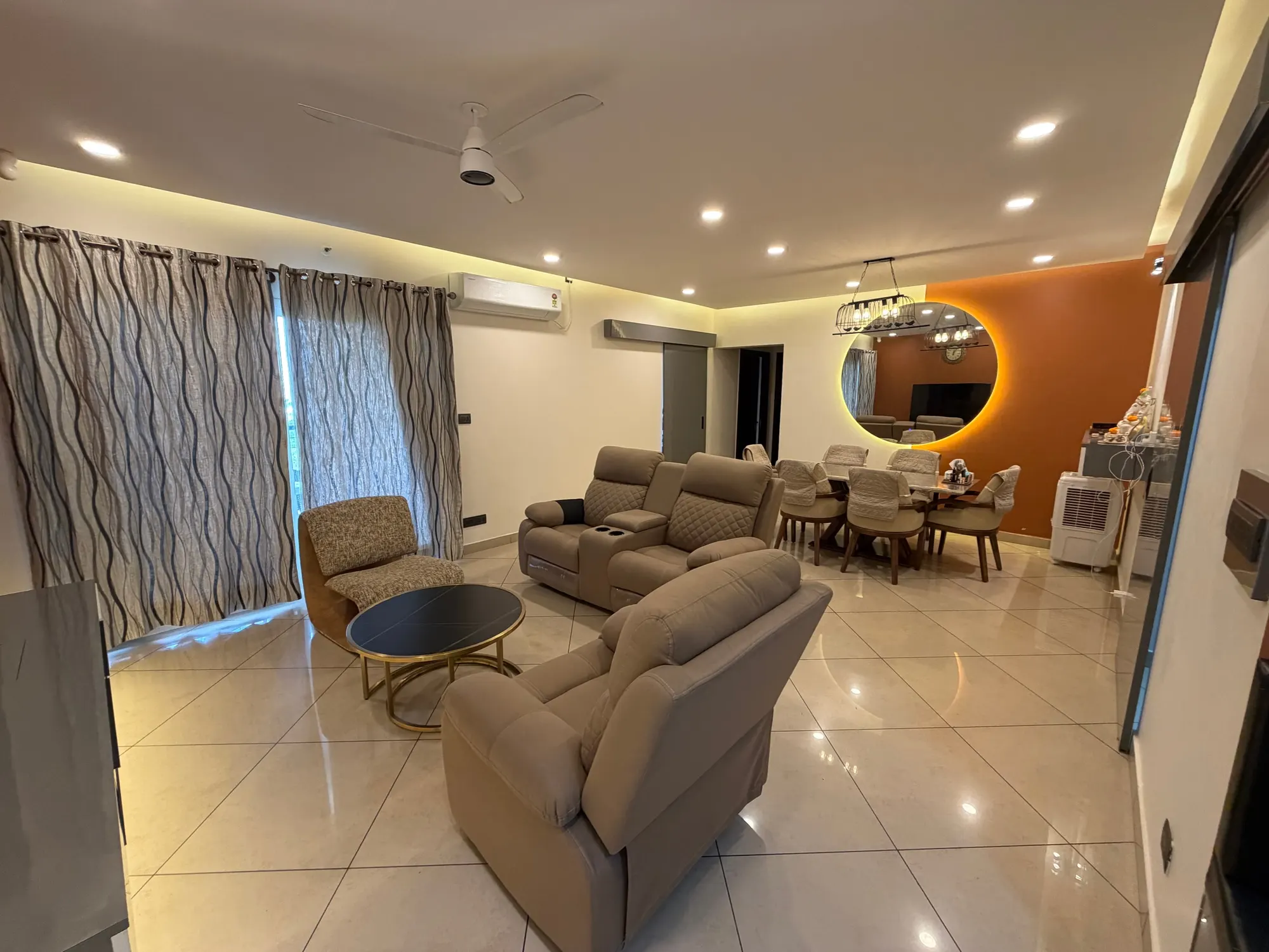 Living and dining space with recliners, a sculpted lounge chair, wave-pattern curtains and a dining table under a cage pendant beside a backlit round mirror