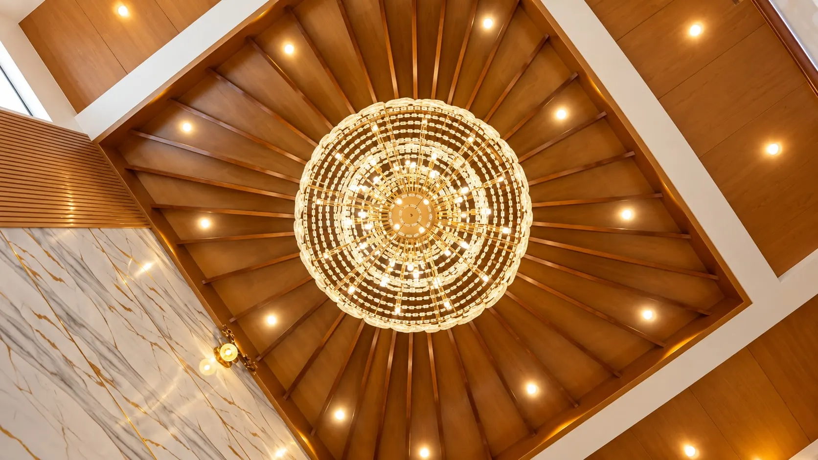 Looking straight up: timber fins radiating from a crystal chandelier, ringed by downlights
