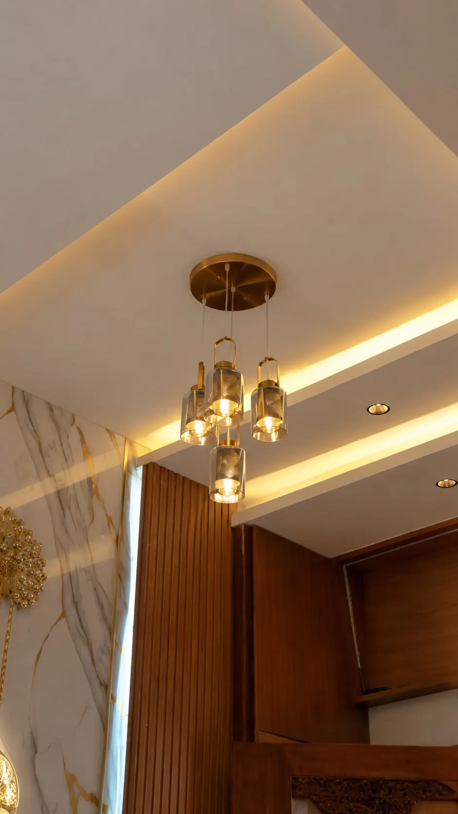 A brass-and-glass cluster pendant beneath a lit ceiling cove, beside a marble-finish wall and timber slats